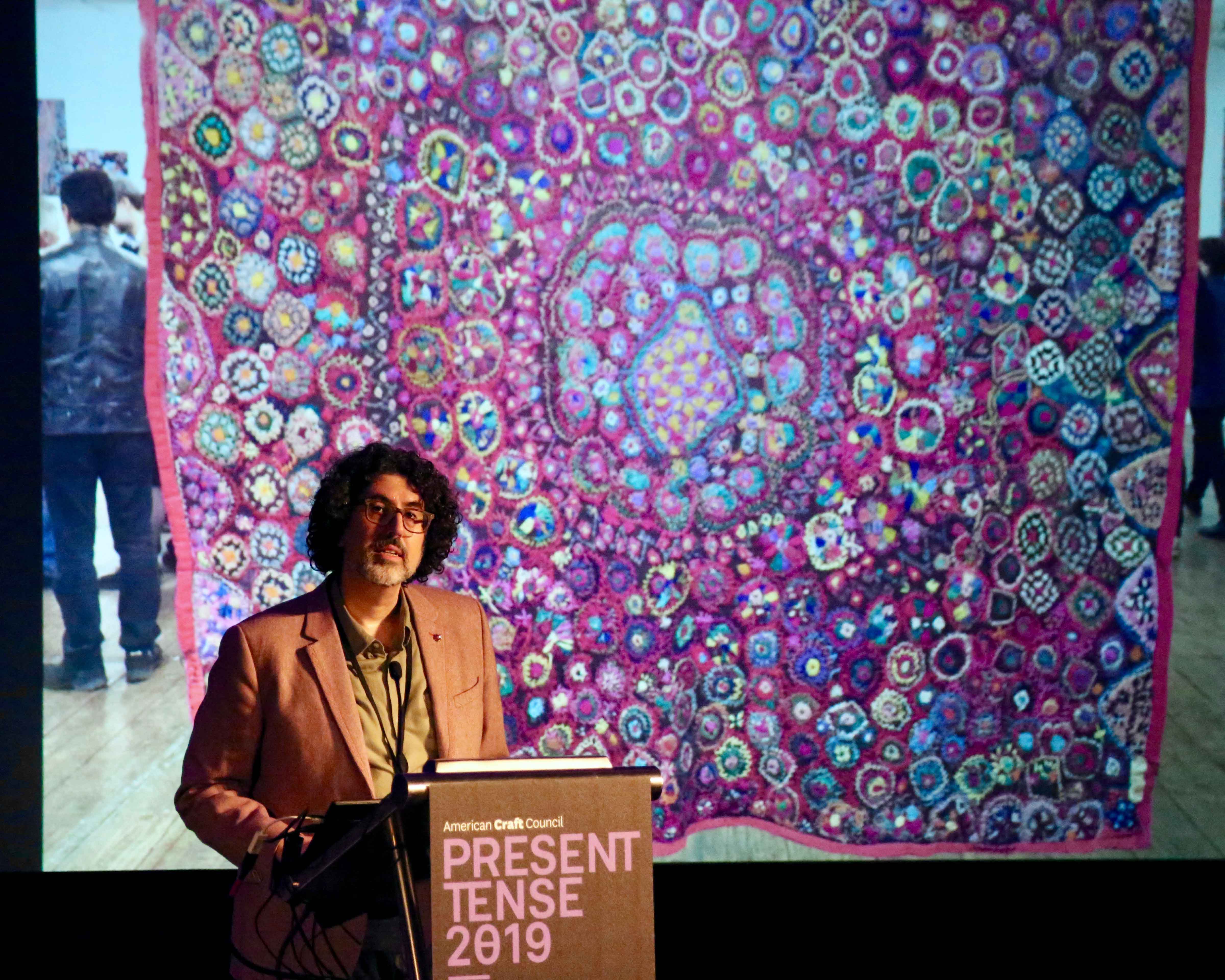 Person speaking at a podium with with an image of a colorful tapestry behind them