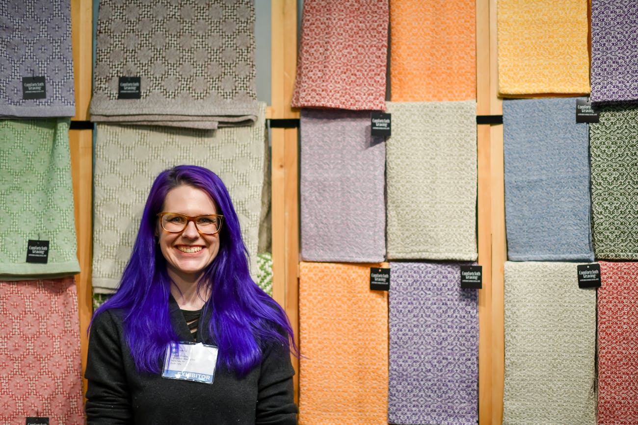 Person with long purple hair smiling in front of wall of handmade linens